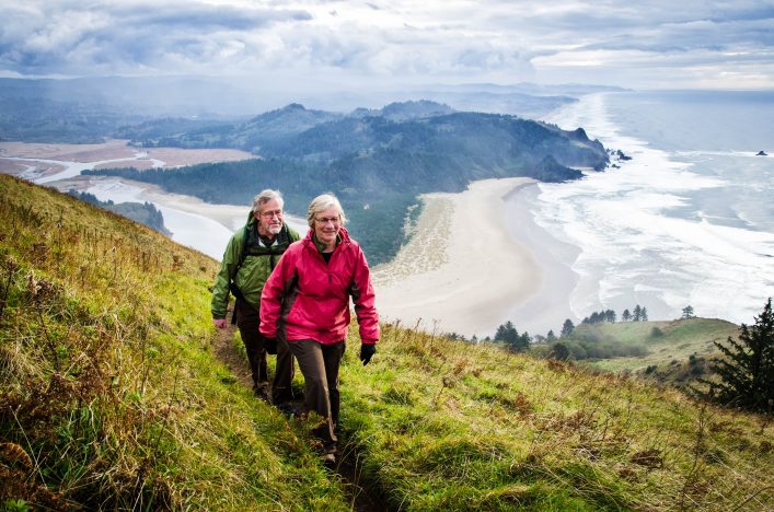 Älteres Paar wandert einen grasbewachsenen Hügel hinauf, links im Vordergrund. Im Hintergrund erstreckt sich eine Küstenlandschaft mit Sandstrand, Meer und bewaldeten Hügeln unter bewölktem Himmel.