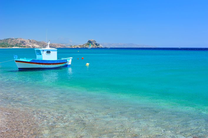 Ein türkisfarbenes Mittelmeer mit einem kleinen, blauen Boot im Vordergrund, das entlang beliebter Kreuzfahrtrouten liegt. Im Hintergrund ist eine felsige Insel zu sehen, darüber ein strahlend blauer Himmel.
