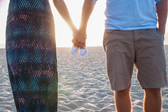 Ein Paar hält sich an den Händen, in der Mitte kleine Babyschuhe. Der Sandstrand und der Sonnenuntergang im Hintergrund schaffen eine romantische Atmosphäre, perfekt für Elternzeit-Reisen.