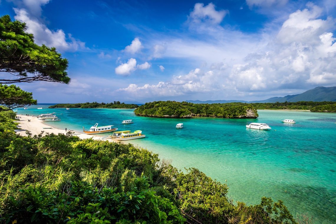 Klares, türkisfarbenes Wasser mit mehreren Booten. Eine üppig bewachsene Insel im Zentrum und ein sandiger Strand links, eingerahmt von grünen Bäumen, unter einem blauen Himmel mit einigen Wolken.