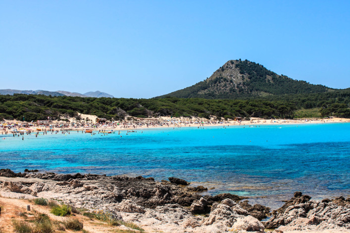 Im Vordergrund Felsen und blaues Meer, dahinter ein belebter Sandstrand. Eine bewaldete Landschaft und ein Berg erstrecken sich im Hintergrund unter klarem, blauem Himmel.