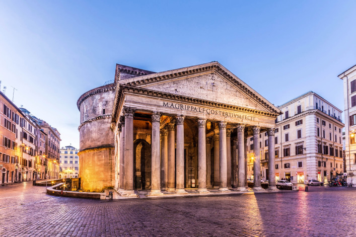 Das Pantheon in Rom bei Dämmerung, frontal aufgenommen. Massive Säulen tragen einen Giebel; dahinter wölbt sich die runde Kuppel. Umgeben von Gebäuden, Kopfsteinpflaster im Vordergrund.