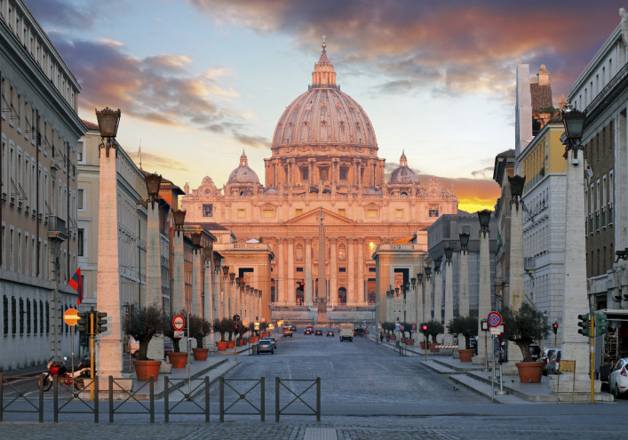 Der Petersdom in Rom, beleuchtet vom warmen Licht des Sonnenuntergangs, thront am Ende einer breiten Straße, die von Gebäuden gesäumt ist. Dramatische Wolken zieren den Himmel.