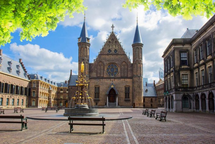 Historisches Gebäude mit markanten Türmen im Hintergrund, im Vordergrund ein Brunnen mit goldener Statue, umgeben von Bänken. Laubbäume rahmen das Bild oben ein, klarer blauer Himmel.
