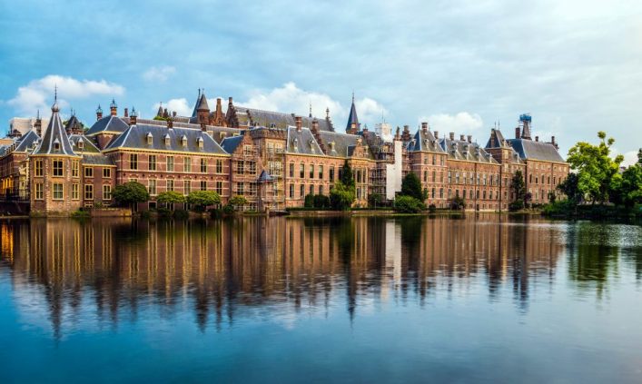 Historische Gebäude des Binnenhofs in Den Haag, Niederlande, spiegeln sich in einem ruhigen Teich. Der Himmel ist bewölkt. Bäume und Büsche säumen den rechten Rand.