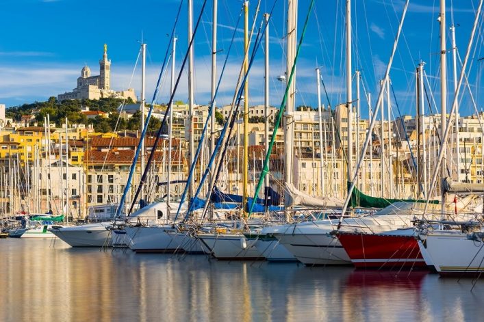 Segelboote im Hafen von Marseille, Frankreich, mit der Basilika Notre-Dame de la Garde auf einem Hügel im Hintergrund. Blaue und grüne Bootsmasten dominieren den Vordergrund, während die Gebäude der Stadt die Mittelachse bestimmen.