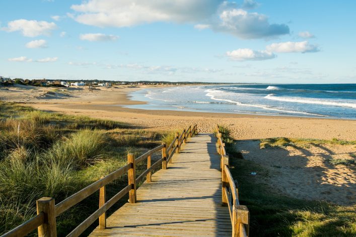 Ein hölzerner Steg führt über eine Dünenlandschaft hinunter zu einem weiten Sandstrand mit sanften Wellen. Im Hintergrund erstreckt sich das Meer unter einem blauen Himmel.