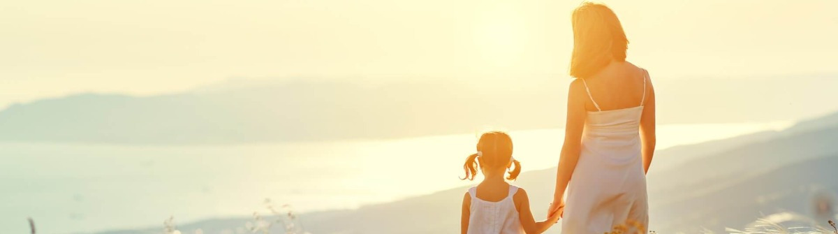 Eine Mutter und ihre Tochter, beide in weißen Kleidern, stehen Hand in Hand auf einem Hügel und blicken auf das Meer. Im Hintergrund sind Berge zu sehen. Sonnenuntergang und sanftes Licht erzeugen eine warme Atmosphäre, ideal für Elternzeit Reisen.