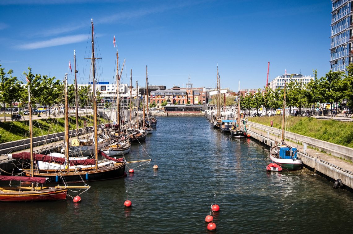 Segelboote liegen an den Kais eines engen Hafenkanals in Kiel, umgeben von modernen Gebäuden und Grünflächen bei strahlend blauem Himmel. An den Kais sind zahlreiche Bäume und rote Bojen auf dem Wasser platziert.
