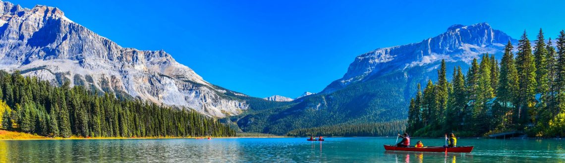 Türkises Wasser, umgeben von Nadelbäumen, mit zwei roten Kanus im Vordergrund. Im Hintergrund erheben sich majestätische Berge unter klarem, blauem Himmel.