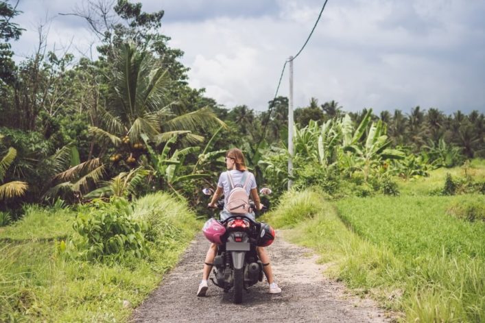 Eine Frau sitzt auf einem Roller auf Bali und steht auf einem unbefestigten Weg. Im Hintergrund sieht man den dichten Wald.