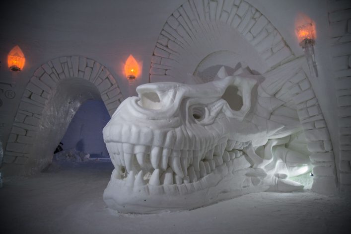 Großer Schneeskulptur eines Drachenkopfes in einem Eishotelraum mit gewölbten Türen. An den Wänden sind Eislaternen angebracht, die warmes Licht spenden. Schneeboden und -wände umgeben die Szene.