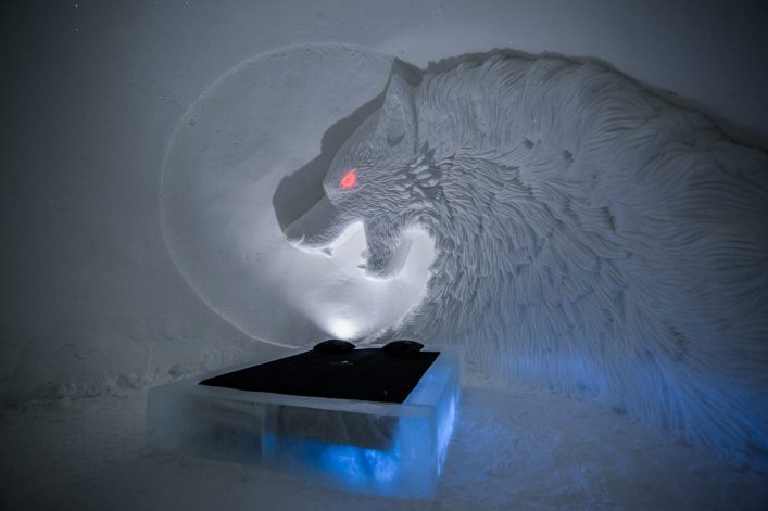 Im Eishotel steht ein Bett aus Eis, darüber eine kunstvolle Wolfs-Skulptur mit leuchtend rotem Auge. Der Wolf ragt aus der rechten Wand hervor, im Hintergrund schimmert blaues Licht.