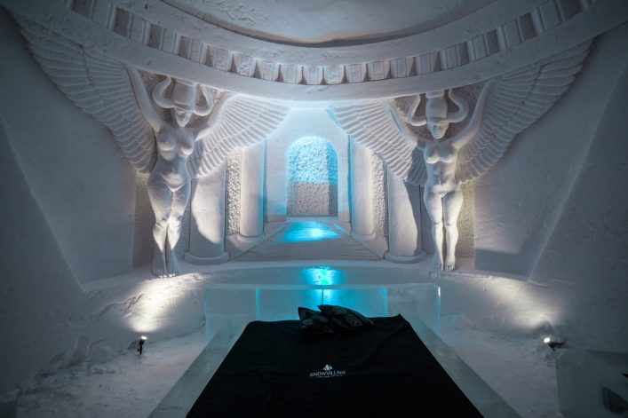 Eisige Skulpturen mit Flügeln zieren die Wände eines Eishotels, flankierend zu einem beleuchteten Gang. Im Vordergrund steht ein Bett aus Eis mit einer dunklen Decke und Kissen.