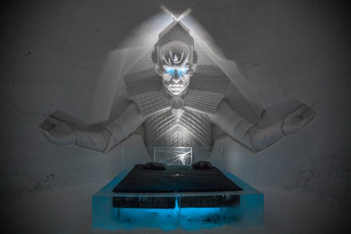 Eiswandrelief in Form einer mystischen Figur mit leuchtend blauen Augen in einem Eishotel. Darunter ein Eisbett mit zwei Kissen, das von blauem Licht erleuchtet wird.