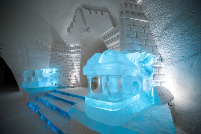 Im Eishotel führen blaue leuchtende Stufen zu zwei detailreichen Eisskulpturen, die wie Drachenköpfe aussehen. Im Hintergrund klare, strukturierte Eiswände mit Ziegel- und Zinnenmustern.