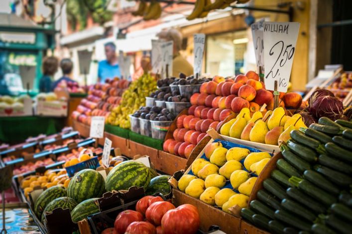 Auf einem Marktstand liegen links Wassermelonen und Tomaten, rechts Berge aus Birnen, Äpfel und Pflaumen. Alle Früchte sind in Holzkisten gestapelt, Preisschilder stecken in den Auslagen.