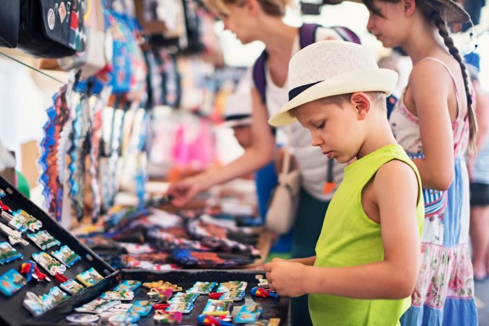 Junge im Vordergrund mit weißem Hut und grünem Shirt betrachtet bunte Magnete auf einem Marktstand; im Hintergrund weitere Personen, die ebenfalls die Waren betrachten. Links sind Taschen und Souvenirs.
