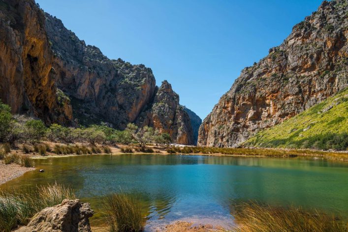 Klares, blaugrünes Wasser eines Sees im Vordergrund, umgeben von hohem Schilf. Im Hintergrund erheben sich steile, felsige Klippen unter blauem Himmel und dichten, grünen Büschen.