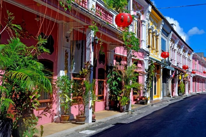 PhuketBunte historische Gebäude in Phuket mit roten Laternen an der Fassade. Palmen und Pflanzen schmücken die schmalen Gehwege. Die Straße verläuft entlang der Häuserfront unter blauem Himmel._Stadt_Thailand