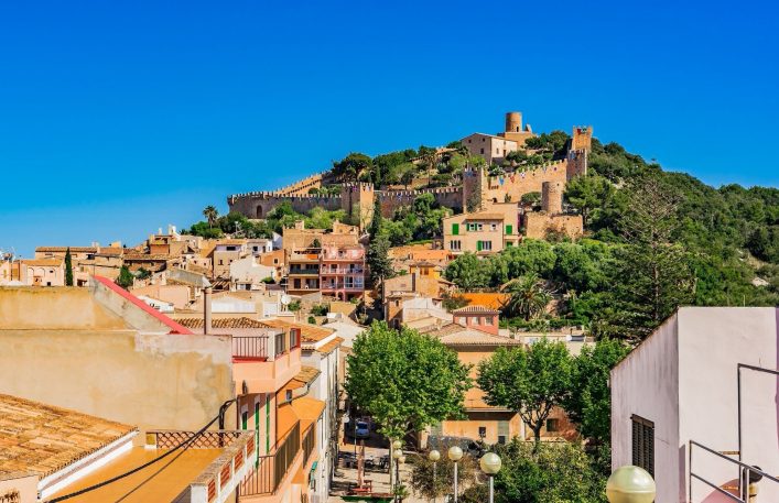 Bunte Häuser drängen sich entlang einer grünen Hügellandschaft. Oben eine mittelalterliche Burg mit Zinnen. Strahlend blauer Himmel überspannt das friedliche Panorama der mediterranen Szene.