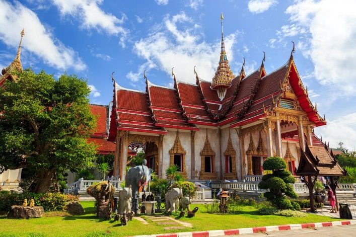 Buddhistischer Tempel in Thailand mit rotem Dach und goldenen Details. Im Vordergrund sind Bäume und Statuen von Elefanten zu sehen. Der Himmel ist blau mit einigen weißen Wolken.