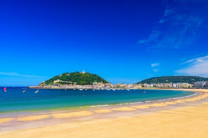 Weißer Sandstrand von La Concha in San Sebastián mit türkisfarbenem Wasser, dahinter ein mit grünen Bäumen bewachsener Hügel. Links und rechts Wohngebäude. Beliebte Kreuzfahrtrouten passieren hier.