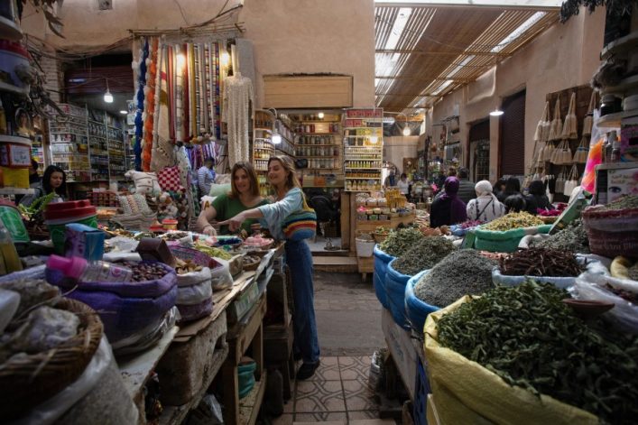In einem lebhaften, überdachten Souk in Marrakesch stehen zwei Frauen vor einem Tisch, der mit bunten Gewürzen und Körnern beladen ist. Links hängen verschiedene Stoffe, im Hintergrund sind weitere Marktstände.