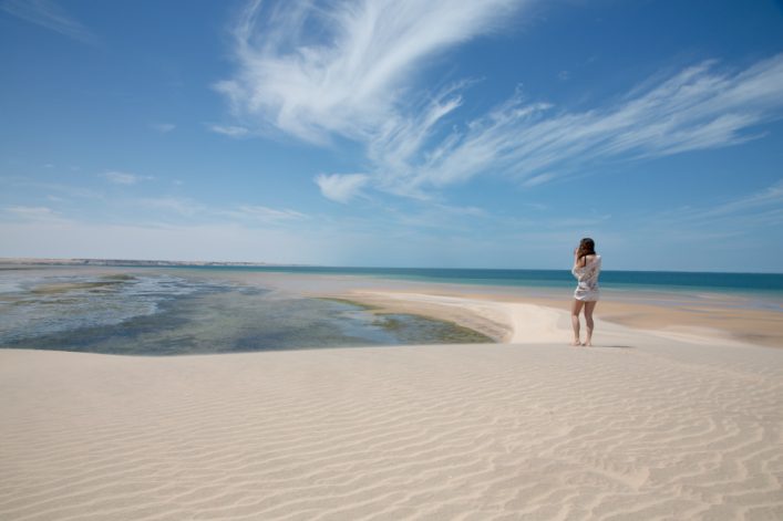 Person steht in der Mitte auf einer Sanddüne und blickt aufs Meer. Rechts der Person, blaues Meer mit klarem Himmel; links und unten, wellenförmiger Sand. Harmonisch fließende Wolken oben.