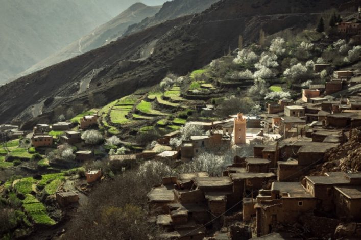 Terrassenförmige Landschaft mit grünen Feldern und blühenden Bäumen, die an Berghängen liegen. Im Vordergrund ein Dorf mit traditionellen Lehmhäusern. Serpentinen führen durch die Berge im Hintergrund.