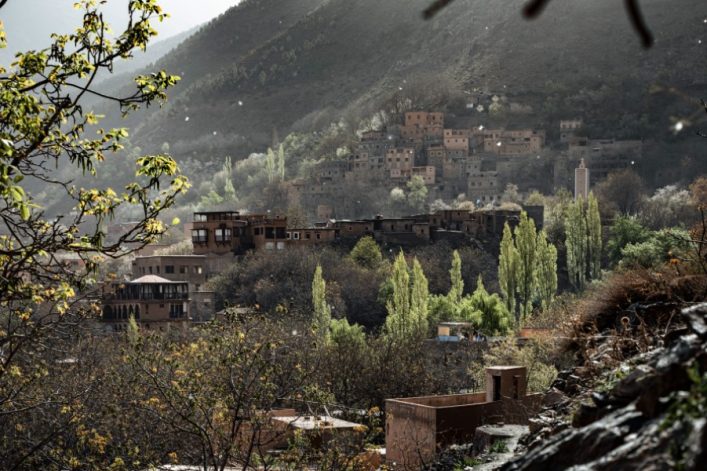 Bergdorf mit traditionellen Häusern am Hang. Im Vordergrund grüne Bäume, vereinzelt blühende Zweige. Im Hintergrund ein bewaldeter Hügel. Vereinzelte Sonnenstrahlen fallen auf das Dorf.