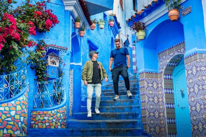 In einer blauen gepflasterten Gasse gehen zwei Männer die Treppe hinab. Links blühen rote Blumen, rechts bunte Fliesen und über ihnen hängen Topfpflanzen. Die Szene wirkt lebendig und farbenfroh.