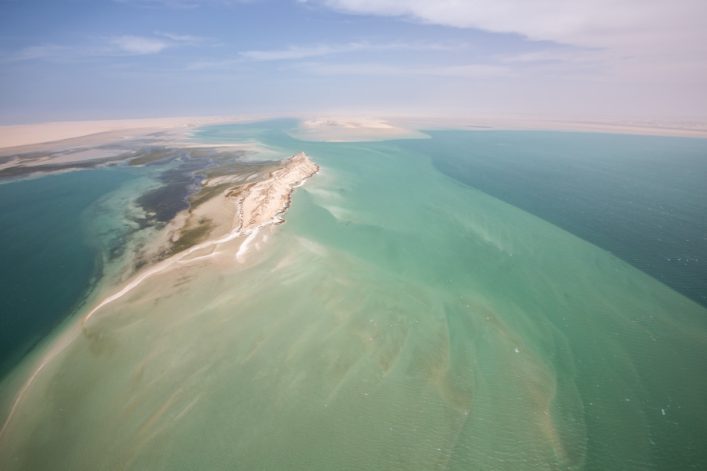 Weitläufige Sandbank erstreckt sich in türkisfarbenem Ozean, von links unten nach rechts oben, umgeben von sich verfärbendem Wasser. Am Horizont ein Himmel mit wenigen Wolken.