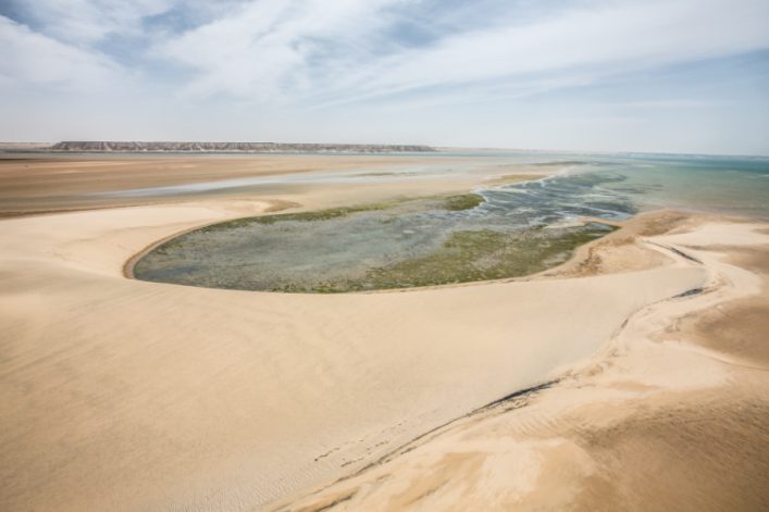 Weite Sanddünenlandschaft mit einer flachen, türkisfarbenen Lagune mittig. Der Himmel ist leicht bewölkt und im Hintergrund erstreckt sich eine Hügelkette.