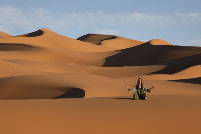 Eine Person sitzt meditierend in der Wüste, umgeben von großen Sanddünen, die sanft geschwungen im Sonnenlicht leuchten. Der Himmel ist blau mit wenigen weißen Wolken.