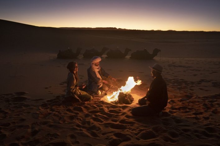 In der Wüste bei Sonnenuntergang sitzen drei Menschen um ein kleines Lagerfeuer. Kamele sind im Hintergrund als dunkle Silhouetten sichtbar, während der Himmel eine orange bis lila Tönung hat.