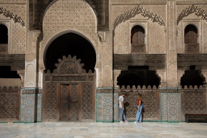 Zwei Personen stehen vor der kunstvoll verzierten Fassade der Medersa Bou Inania in Fès mit Mosaiken und geschnitzten Holzelementen. Drei bogenförmige Fenster oben und eine große Holztür in der Mitte.