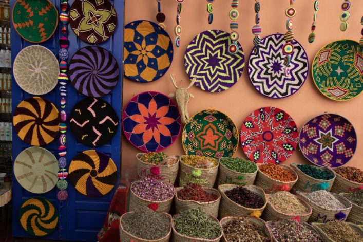 Bunte Flechtkörbe mit geometrischen Mustern hängen an einer Wand in einem Souk. Darunter stehen Säcke mit verschiedenen getrockneten Kräutern und Gewürzen in leuchtenden Farben.