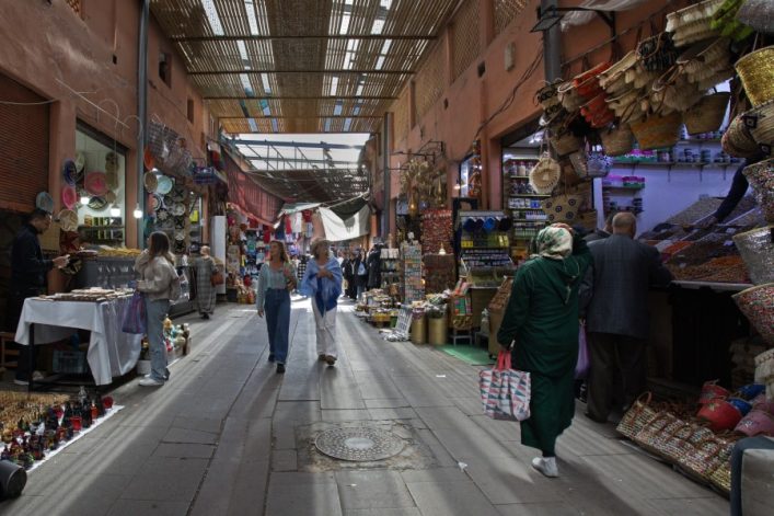 Ein belebter Souk mit überdachtem Gang. Links Verkaufsstände mit bunten Tellern, rechts Körbe und Gewürzberge. Menschen flanieren, darunter eine Frau mit Kopftuch und gestreifter Tasche.