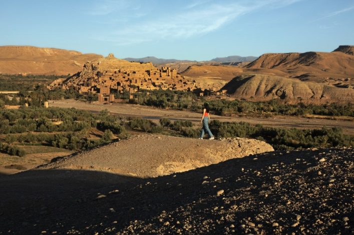Person in Jeans und rotem Oberteil geht auf einem Hügel. Im Hintergrund sind die Lehmbauten von Ait Ben Haddou zu sehen, umgeben von grüner Vegetation und Bergen unter einem blauen Himmel.