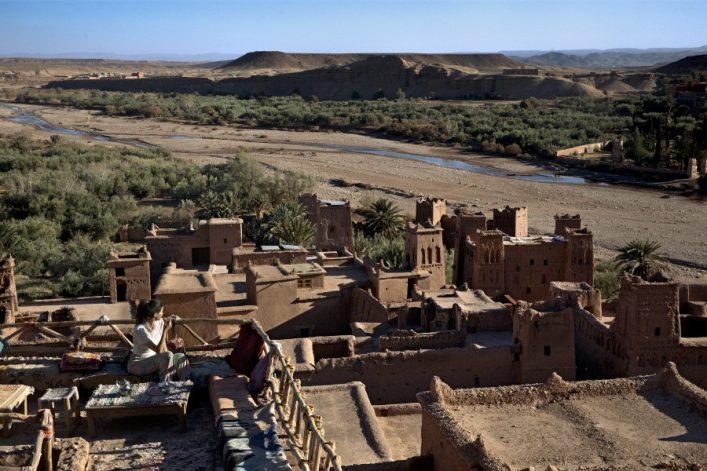 Historisches Lehmdorf in Wüstenlandschaft; im Vordergrund eine Terrasse mit einer Frau; im Hintergrund Palmen, ein Flussbett und Berge.