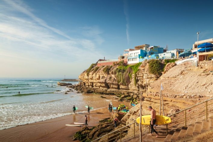 Surfer mit Boards am Strand vor felsiger Küste; oben bunte Häuser in Hügeln, im Hintergrund Meer und ein klarer Himmel; Personen gehen eine Treppe hinunter zur rechten Seite.