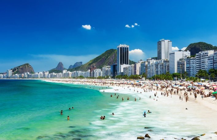 Die Copacabana in Rio de Janeiro an einem sonnigen Tag. Links das türkisfarbene Meer mit Schwimmern. Rechts der breite, weiße Sandstrand voller Menschen und bunten Sonnenschirmen. Im Hintergrund Hochhäuser und grüne Hügel. Die Copacabana ist eine beliebte Station auf Kreuzfahrtrouten durch Südamerika.