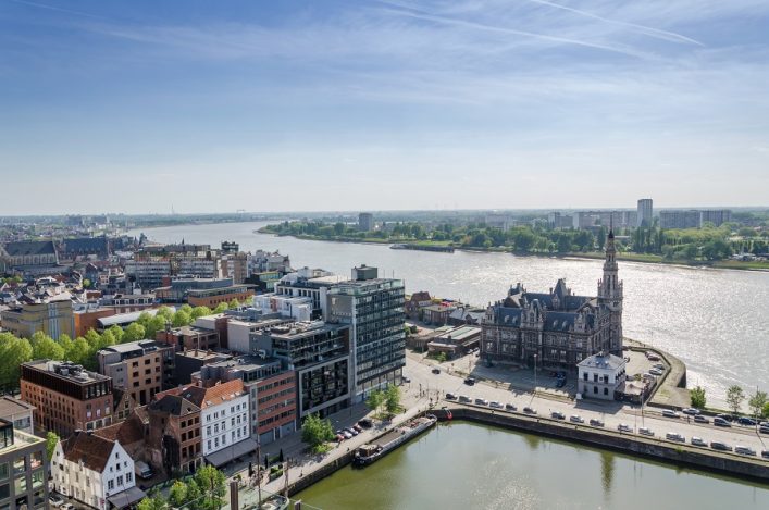 Panoramaansicht des Hafens von Antwerpen: Im Vordergrund moderne und historische Gebäude, dahinter ein Fluss, der unter blauem Himmel glitzert. Rechts prägt ein imposantes Bauwerk die Szene.