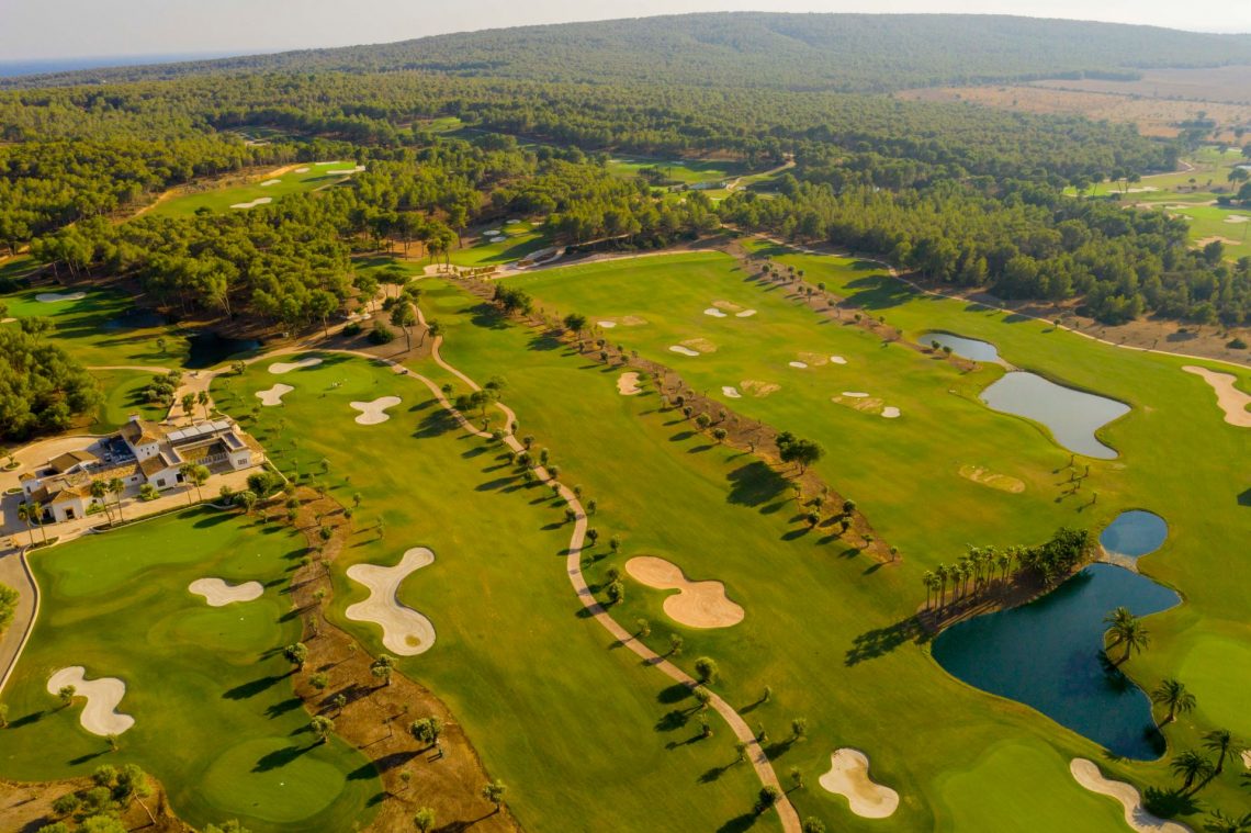 Grüne Golfplatzlandschaft auf Mallorca mit flachen, geschwungenen Sandbunkern und Teichen. Im Vordergrund ein Clubhaus, im Hintergrund bewaldete Hügel; das Ganze bei Sonnenuntergang.