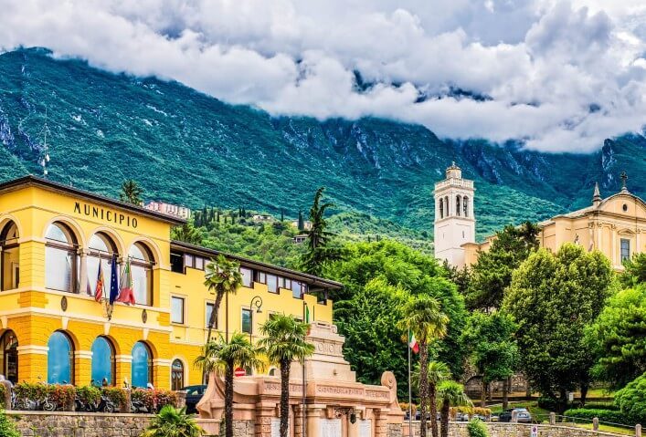 Blick auf die Berge, die die Stadt umrahmen. Die Wolken hängen tief über den Berspitzen. Die Berge selbst sind grün bewachsen. Am rechten Bildrand läasst sich eine imposante Kirche in hellem Stein erkennen. Am linken Rand sticht ein gelb leuchtendes Gebäude ins Auge, das die Aufschrift Municipo trägt. Dazwischen stehen viele grüne Bäume und Büsche.