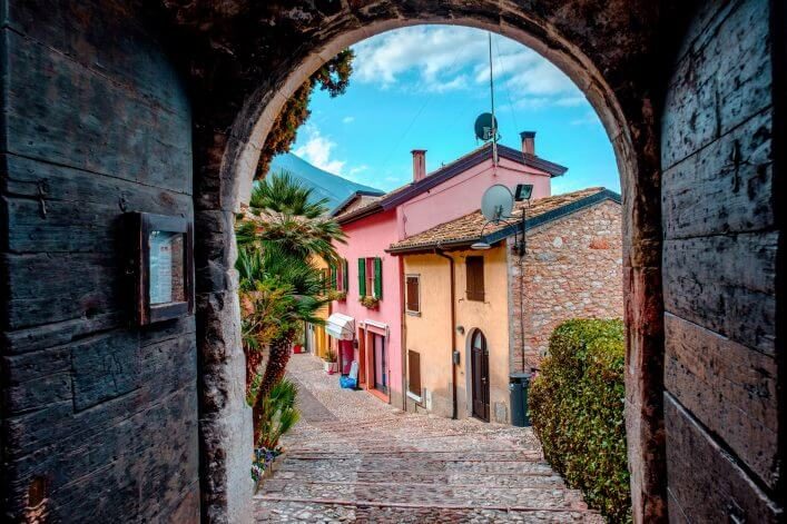 Gasse in Malcesine am Gardasee. Das Foto wurde in einem mit Holz verkleideten Tunnel aufgenommen und nach dem runden Bogen am Ende lassen sich die bunten Häuschen in der Gasse erkennen. Sie sind orange und rosa mit grünen Fensterläden.