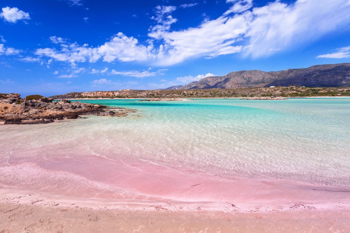 Rosa eingefärbter Strand Elafonissi im Südwesten Kretas: in rosafarbener Sandstrand geht in türkisfarbenes Meer über. Ideal für einen Strandurlaub. Im Hintergrund Berge und blauer Himmel mit wenigen Wolken.