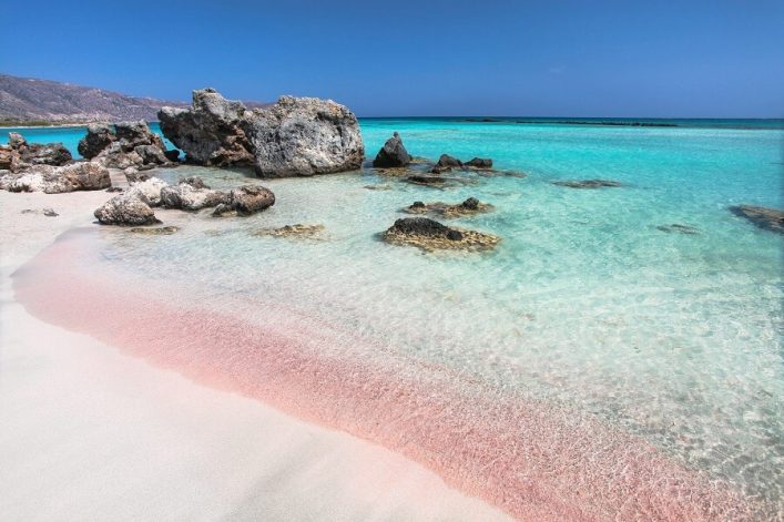 Sanfte Wellen treffen auf rosa Sand an einem Strand mit klarem türkisfarbenem Wasser. Im Hintergrund erheben sich felsige Klippen, die einen idyllischen Küstenabschnitt auf Kreta bilden.