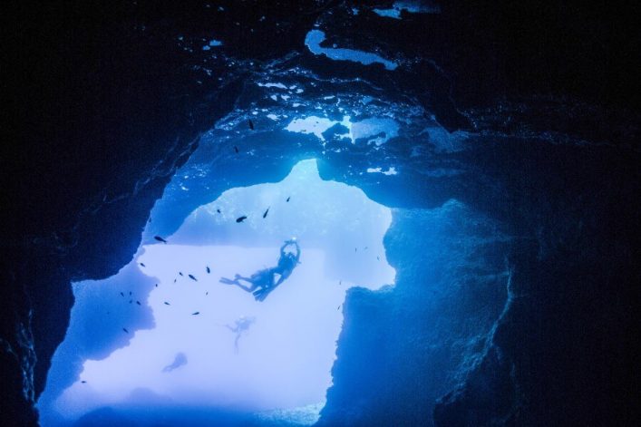 In einer blauen, von Felsen umrahmten Unterwasserhöhle schwimmen mehrere Taucher. Licht strömt von oben herab, während kleine Fische umherziehen. Eine mystische, stille Szenerie.